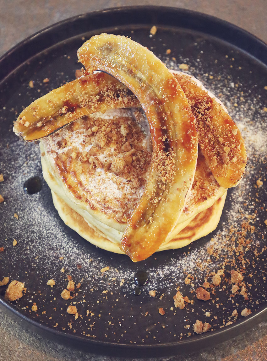Stack of fluffy pancakes topped with caramelised banana, biscuit crumb and a dusting of icing sugar, served on a dark plate at ICE restaurant in Bury St Edmunds, Suffolk.