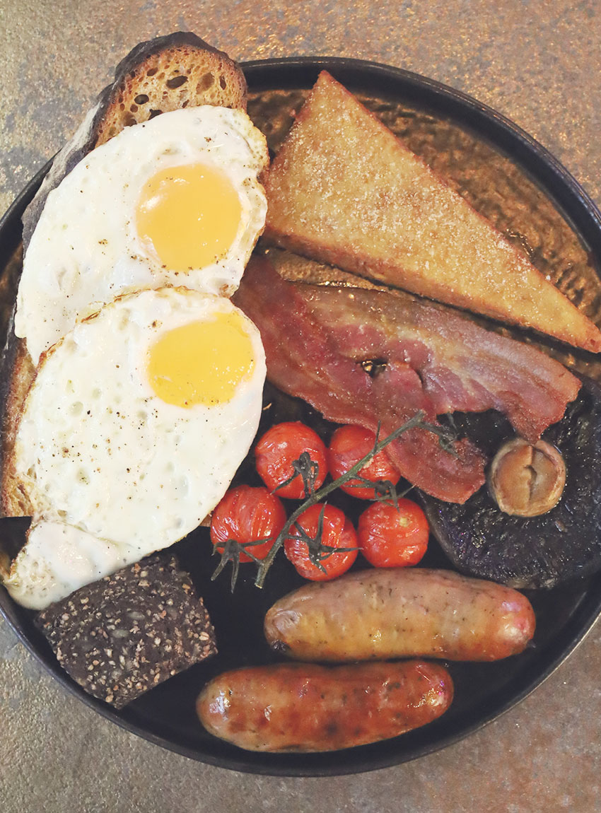 Full English breakfast served at ICE restaurant in Bury St Edmunds, Suffolk, featuring two fried eggs, grilled bacon, sausages, black pudding, roasted vine tomatoes, grilled mushroom, hashbrown, and toasted sourdough on a dark plate.